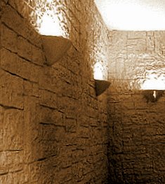 Wnętrze łazienki wykończone płytkami imitującymi kamień w ciepłej, brązowej tonacji. Na ścianach zamontowane trójkątne półki z wbudowanym oświetleniem punktowym, tworzące nastrojowy klimat.